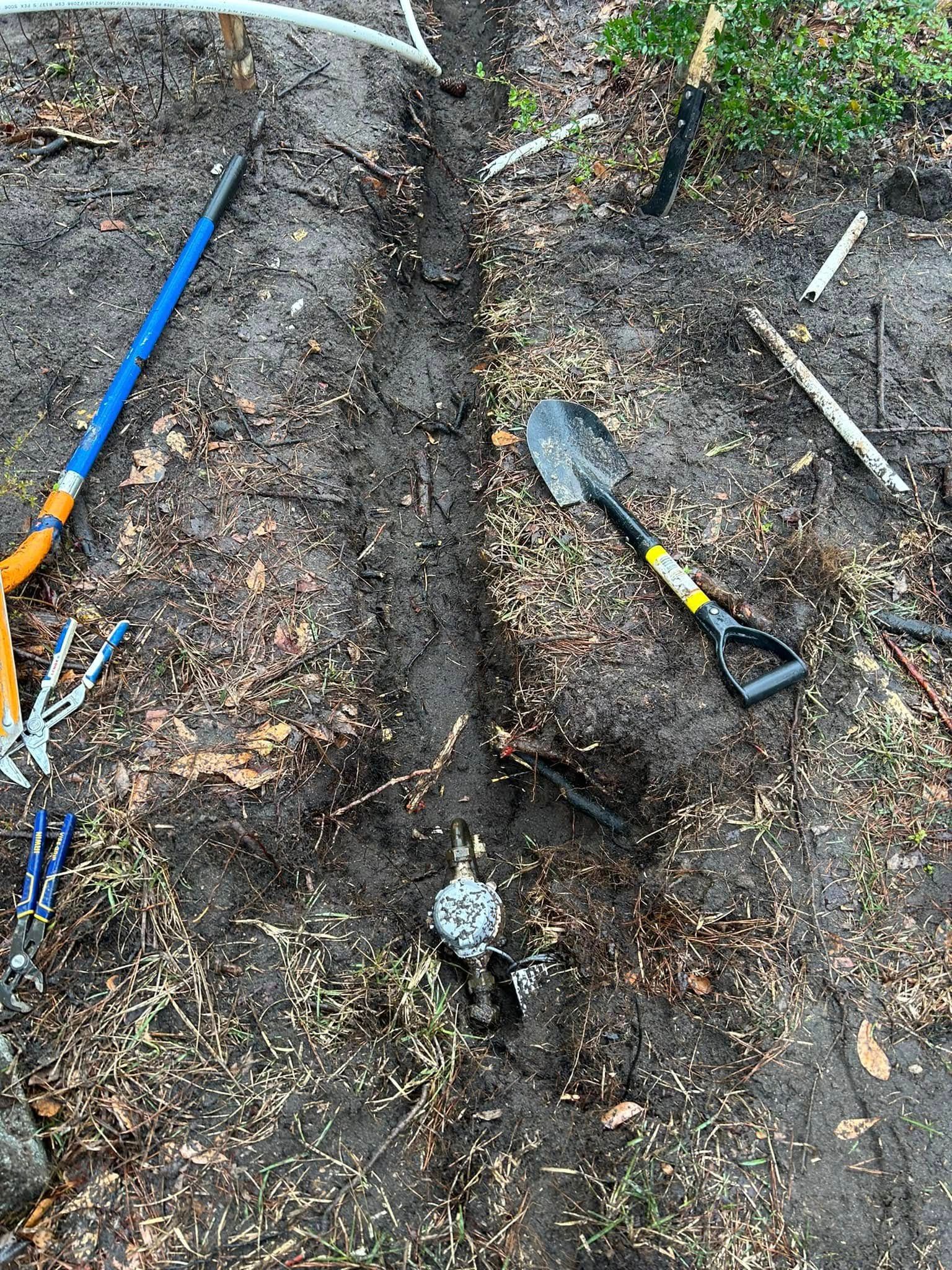 A shallow trench dug in dirt with a shovel, pruning shears, and a pipe repair fitting visible on the ground.