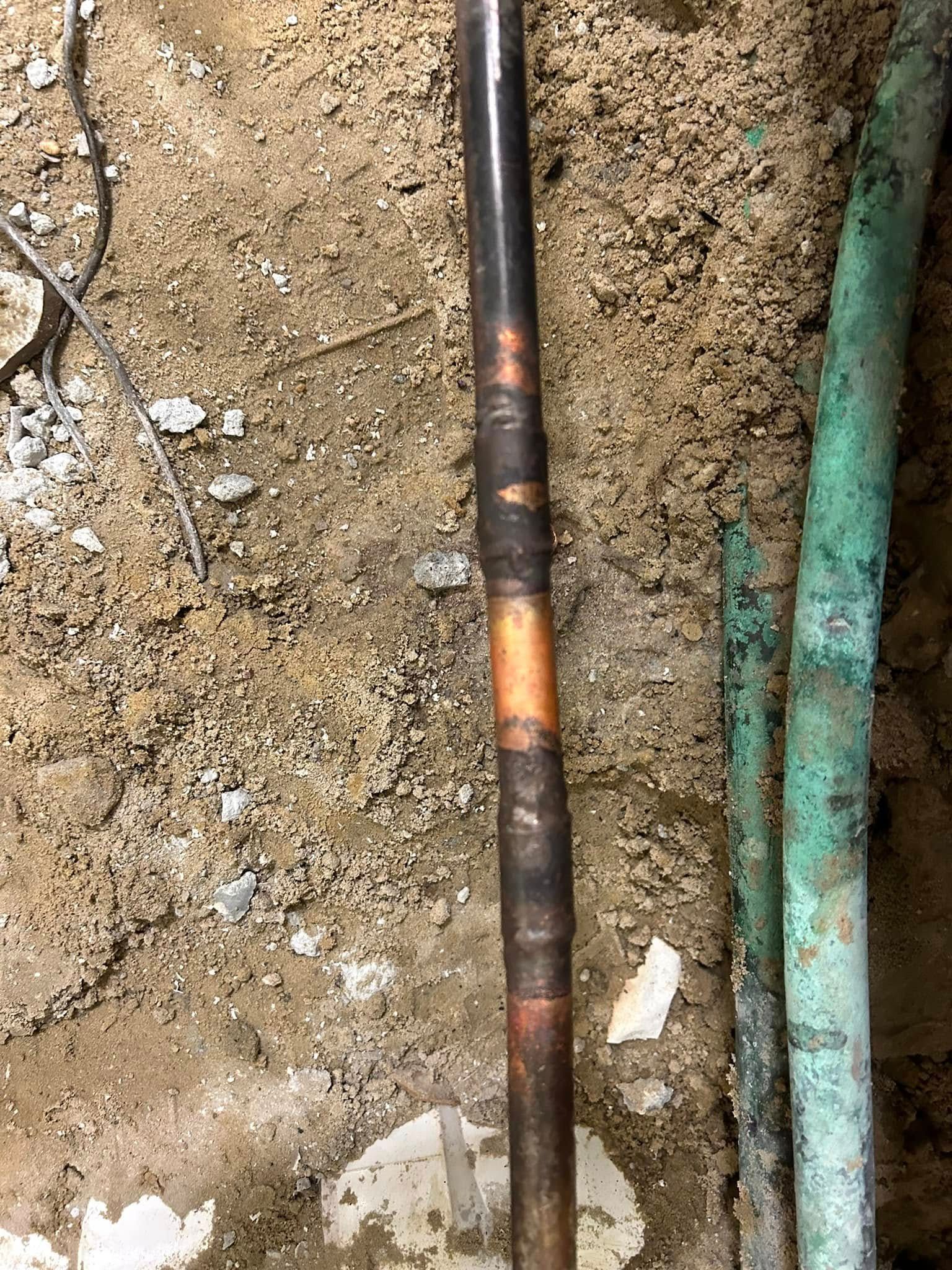 A copper pipe with a soldered repair joint lies on dirt, next to two weathered, green-tinted pipes.
