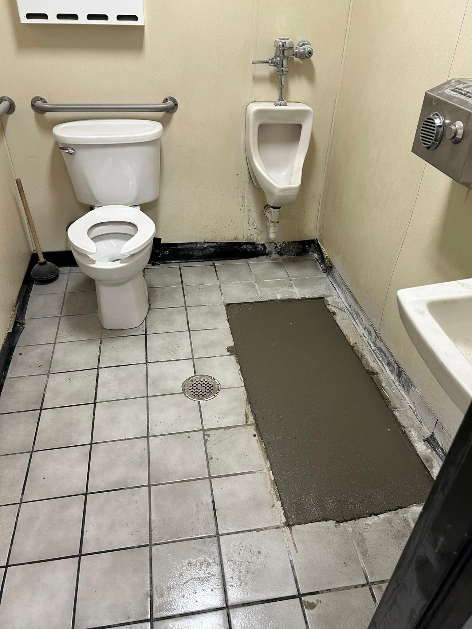 A public restroom with a white toilet and urinal, a floor drain, and a patch of wet concrete on the tiled floor.