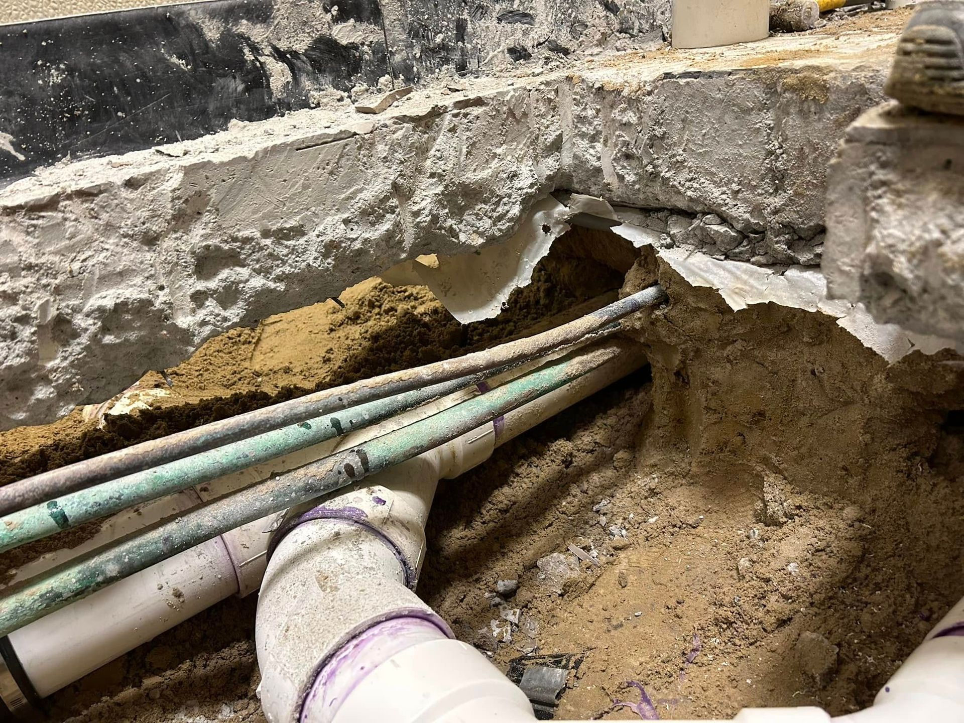 Construction site showing underground pipes and wiring exposed in a trench below a broken concrete floor.