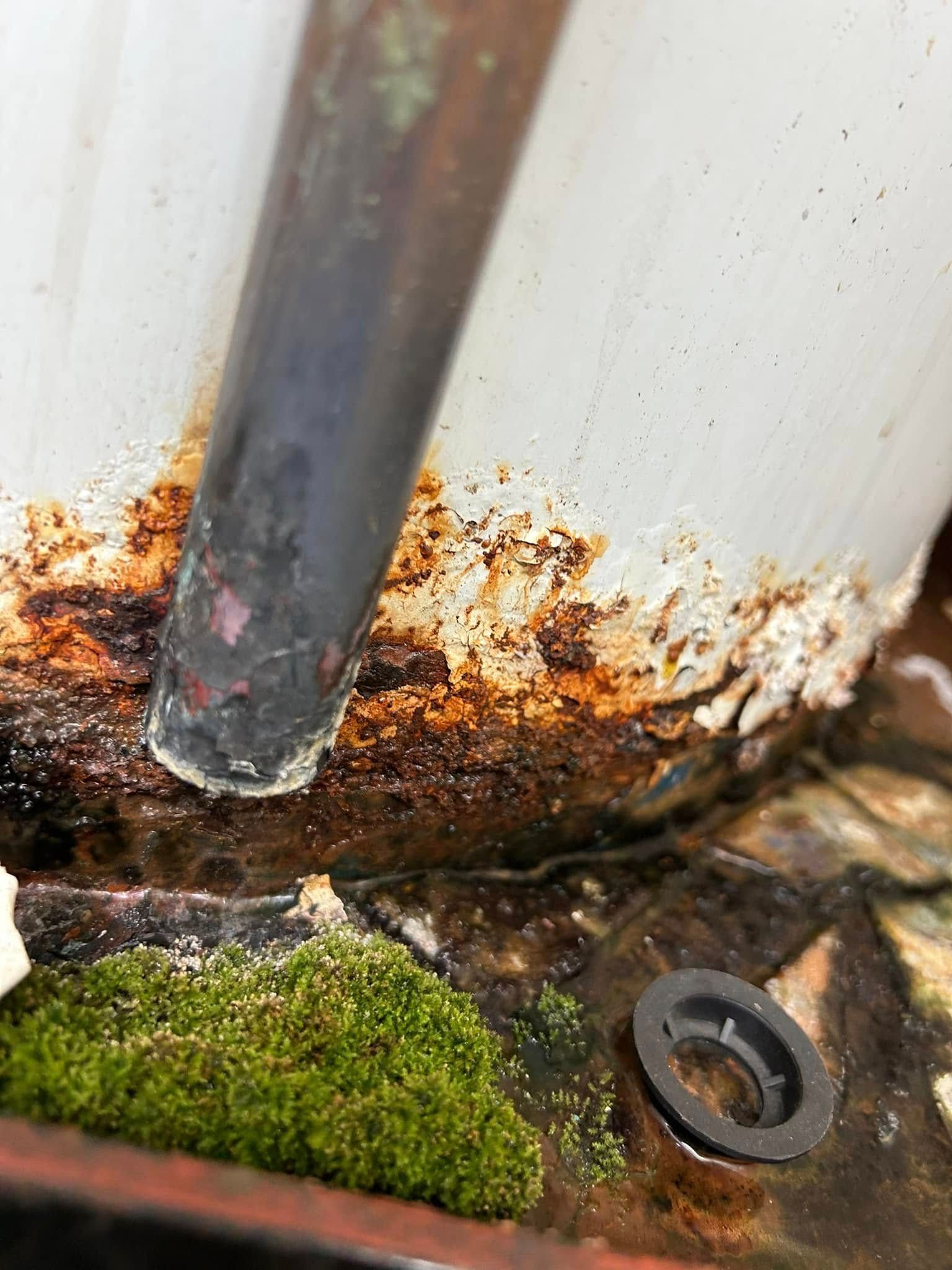 A vertical pipe meeting a rusted base covered in mineral deposits and a patch of green moss.