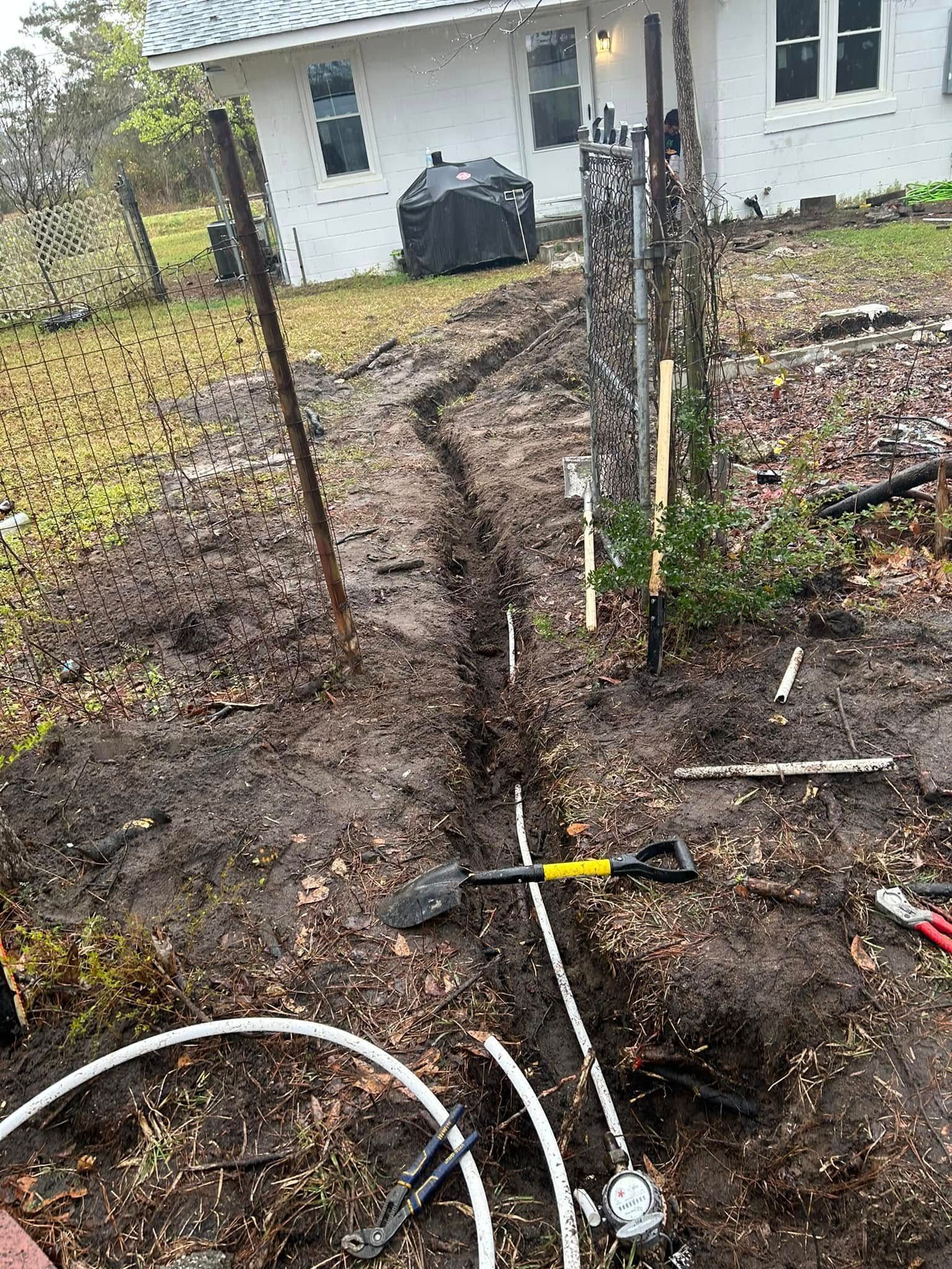 A narrow trench containing a white pipe leads toward a house, with a shovel and tools lying in the dirt nearby.
