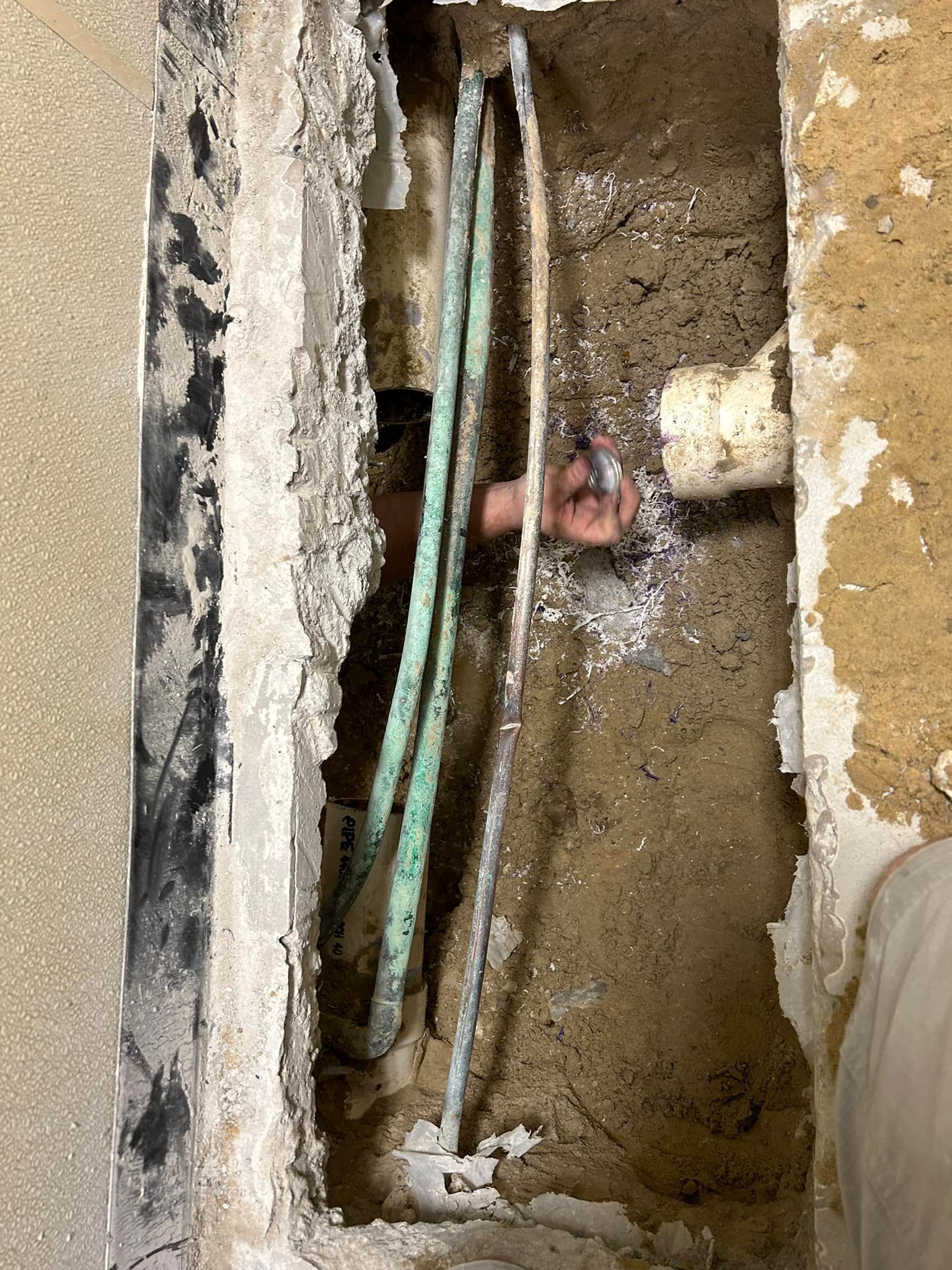 A worker’s hand reaches into an open trench in a concrete floor, surrounded by exposed copper pipes and a white PVC pipe.