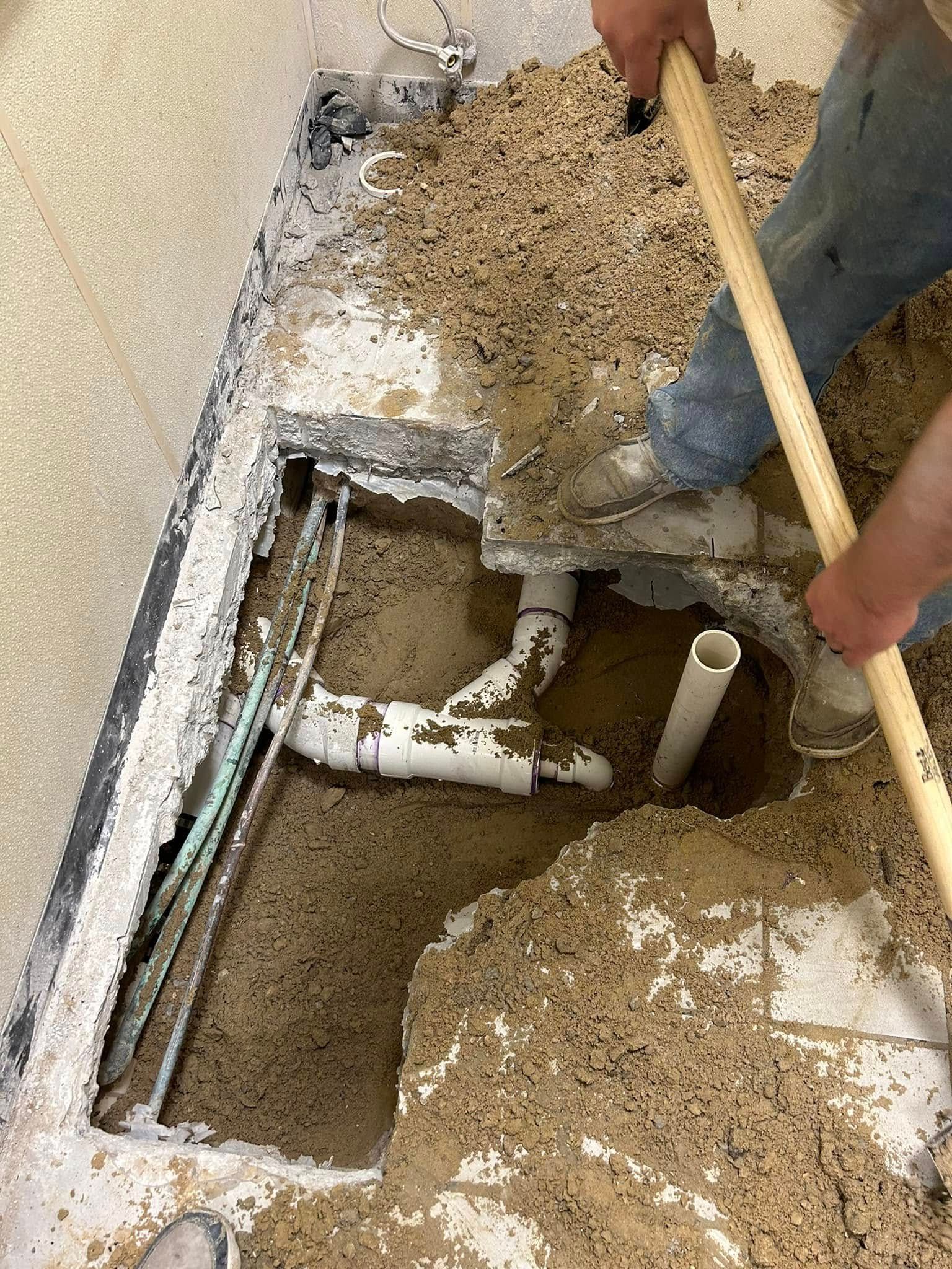 A worker uses a shovel to clear dirt from a concrete floor cutout revealing white PVC plumbing pipes.