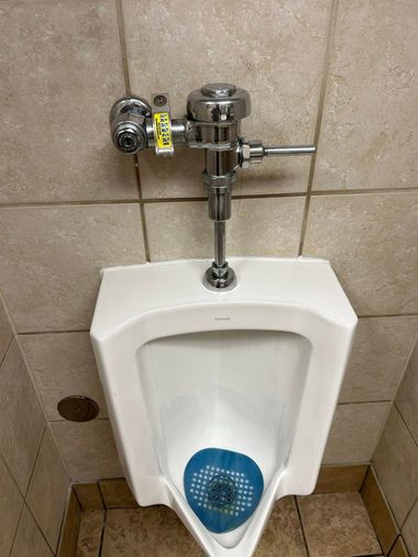 A white wall-mounted urinal with a chrome flush handle and a blue urinal screen in a tiled restroom.