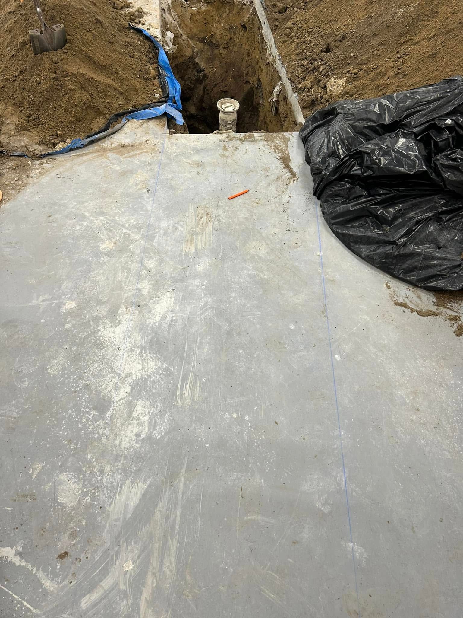 A concrete surface with a dirt-filled trench containing a white pipe, with blue plastic and a black tarp nearby.