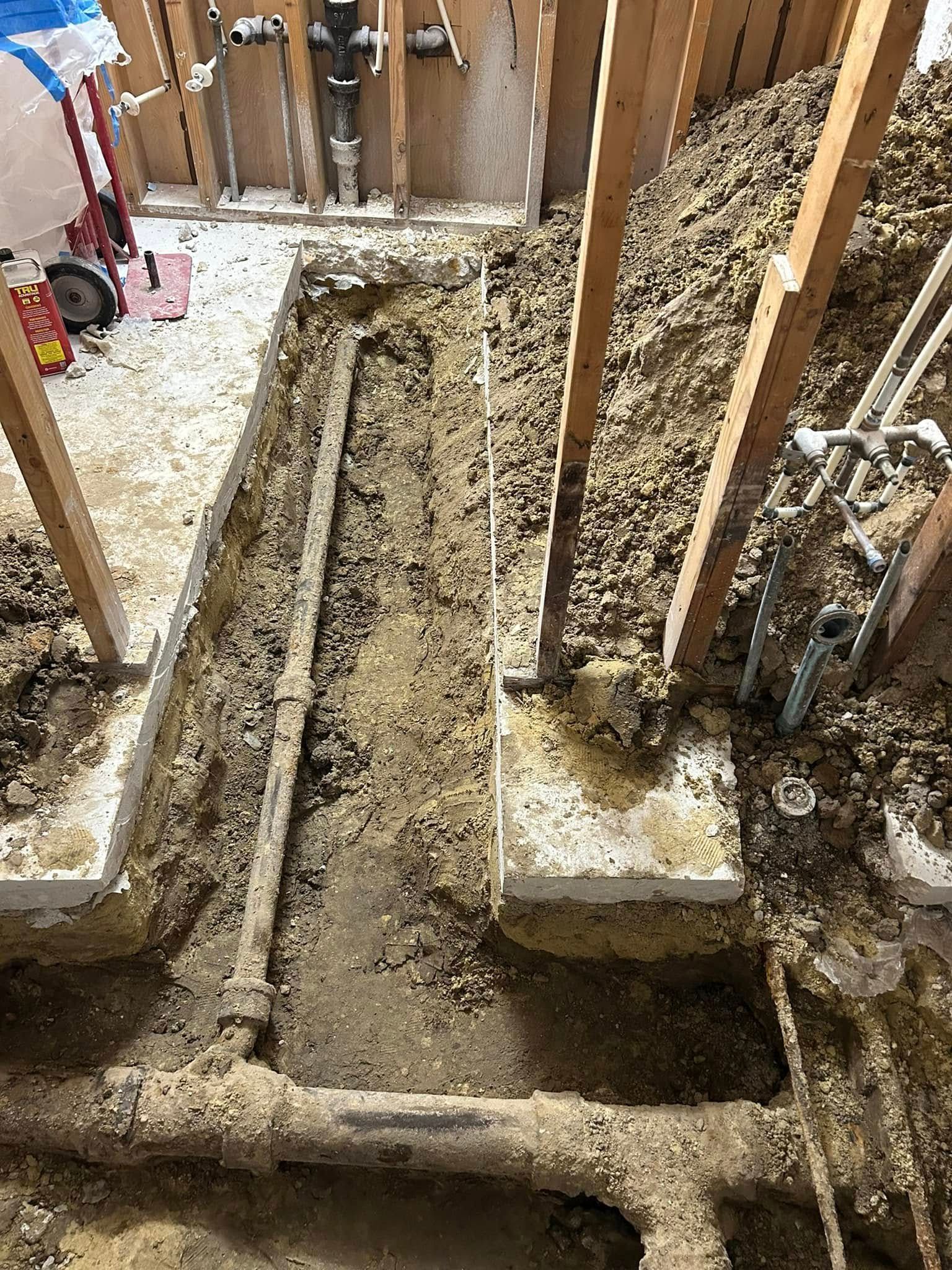 Exposed dirt trench with metal plumbing pipes running through an unfinished room under construction with wall studs.