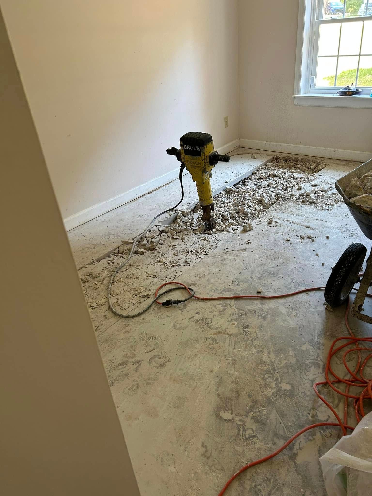 A jackhammer stands in a room with a partially removed floor, leaving concrete debris scattered across the floor surface.