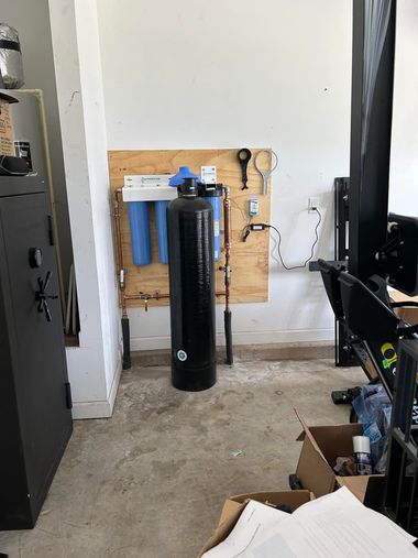 Water filtration system with a large black tank and blue filter housings mounted on a plywood board in a garage.
