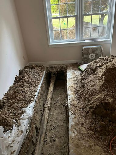 A trench cut into a residential concrete floor, exposing underground pipes, with piles of excavated dirt on either side.