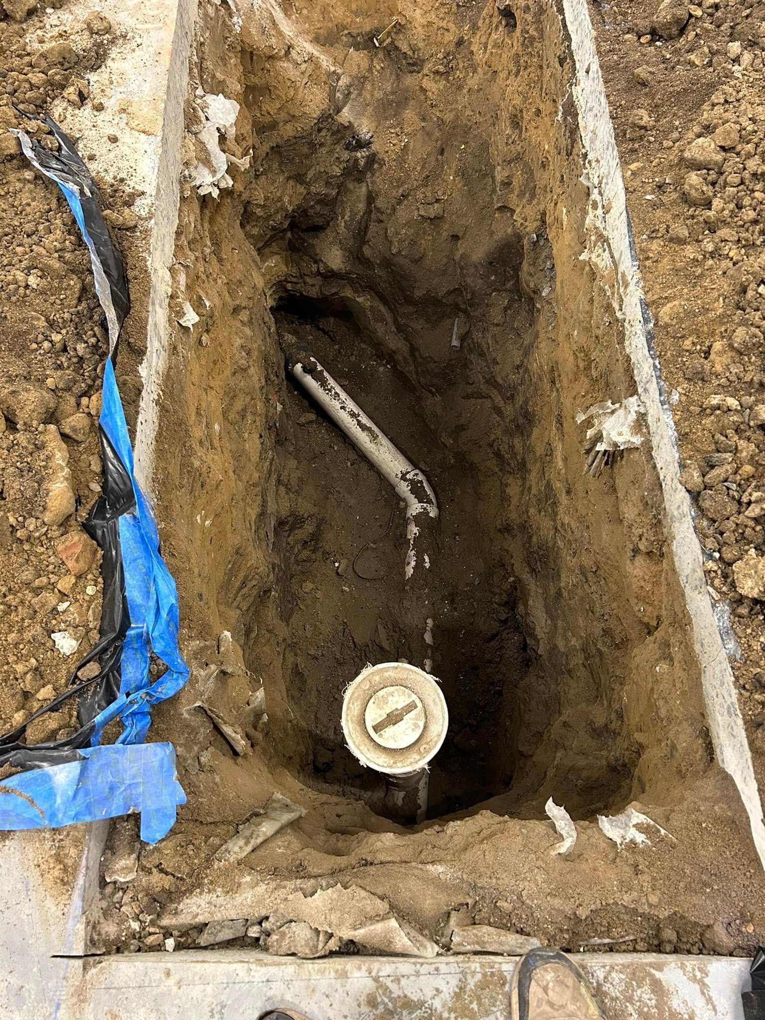 A vertical view of an excavation trench showing a white circular pipe cap and a diagonal grey pipe section in brown dirt.