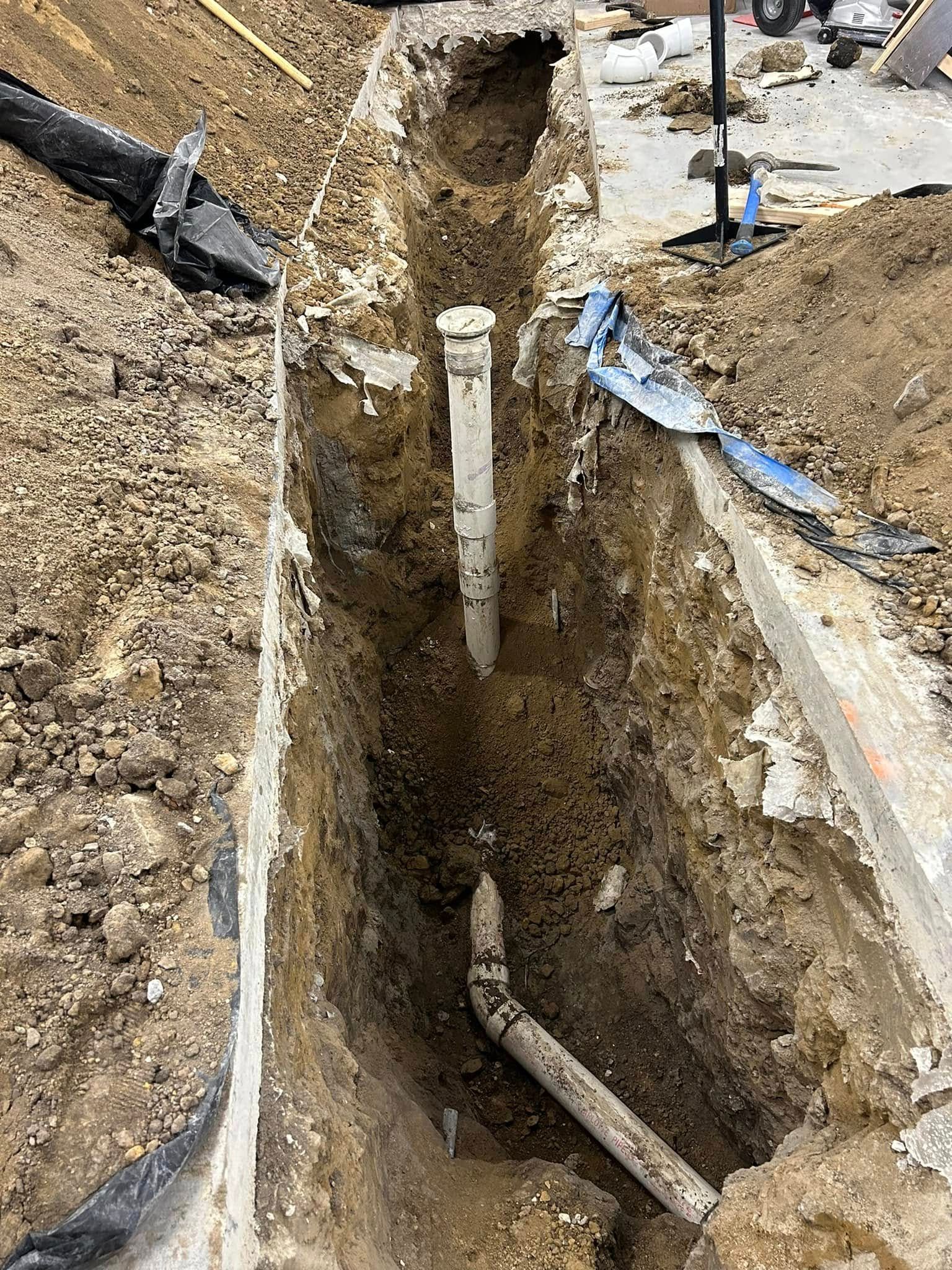 An open trench at a construction site showing white plumbing pipes installed in the dirt.
