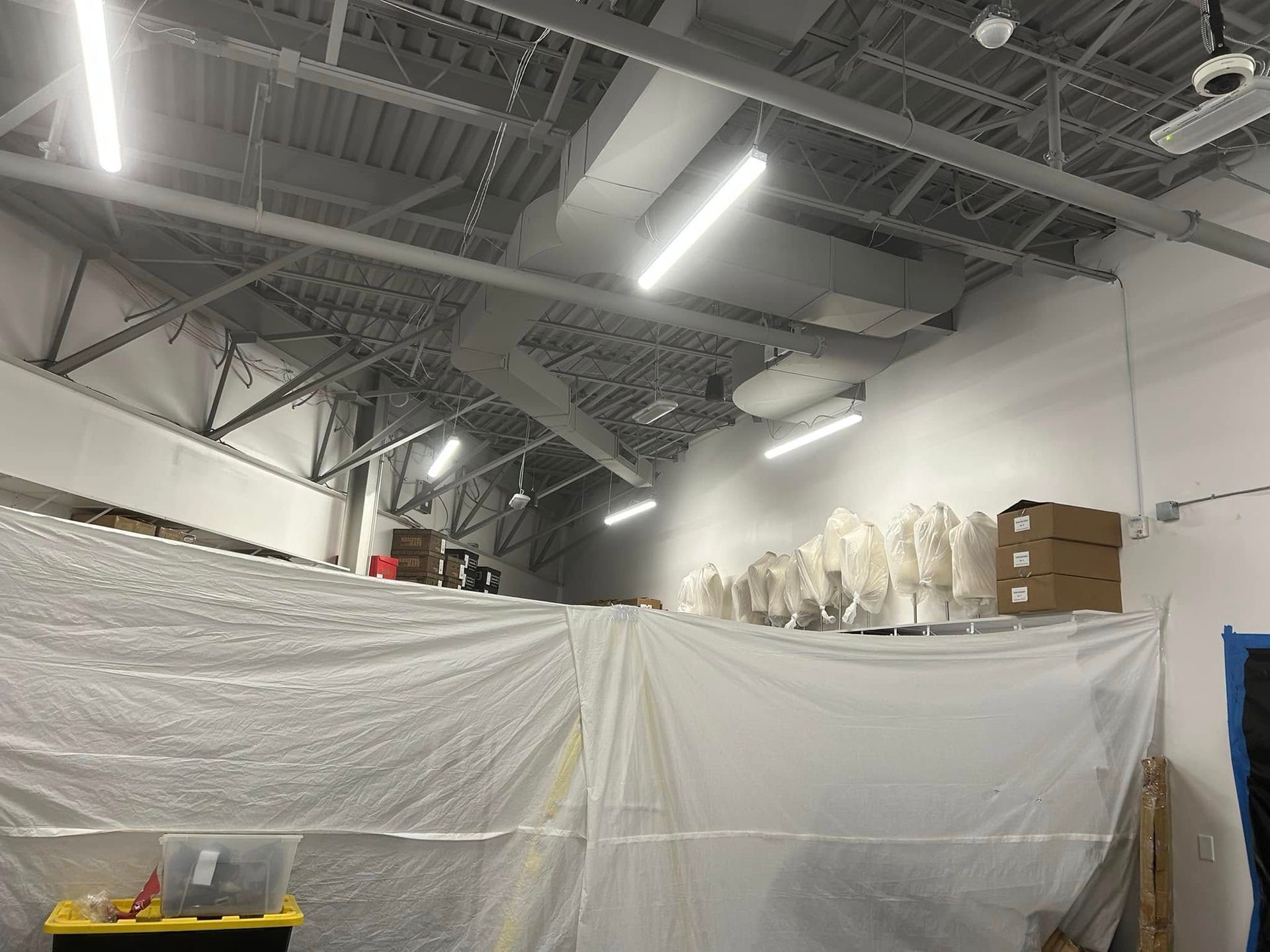 Industrial space with a plastic sheet partition, exposed ceiling, overhead lights, and storage boxes on a high shelf.