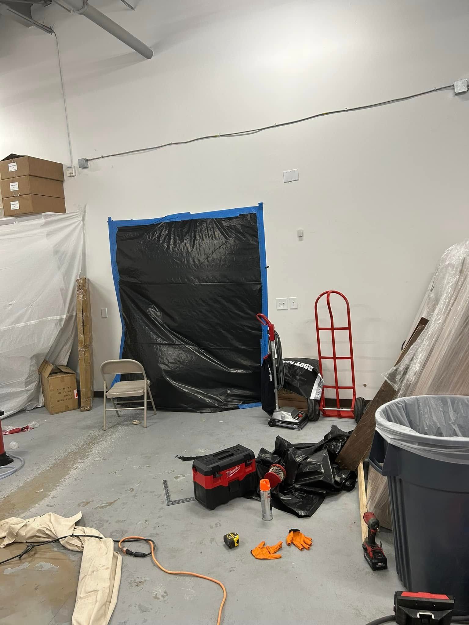 A workspace showing a taped-off door covered in black plastic, a red hand truck, scattered tools, and a trash bin.