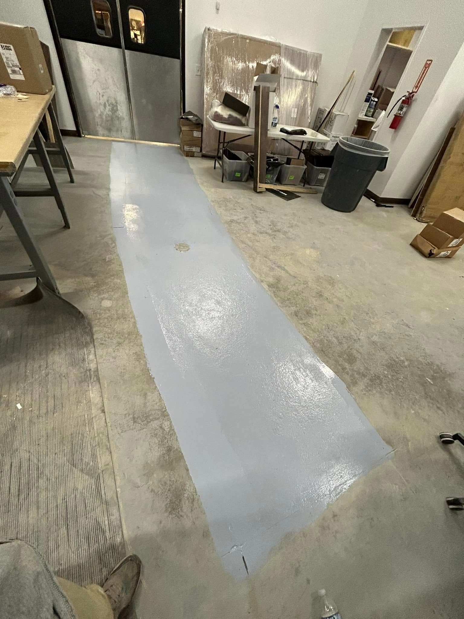A wide, rectangular strip of light gray, glossy floor coating applied to a concrete floor in a room or workshop.