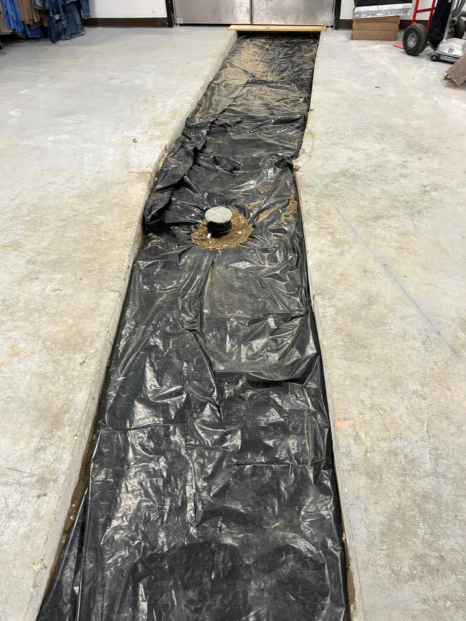 A concrete floor trench lined with black plastic sheeting, featuring a central pipe fitting for plumbing work.