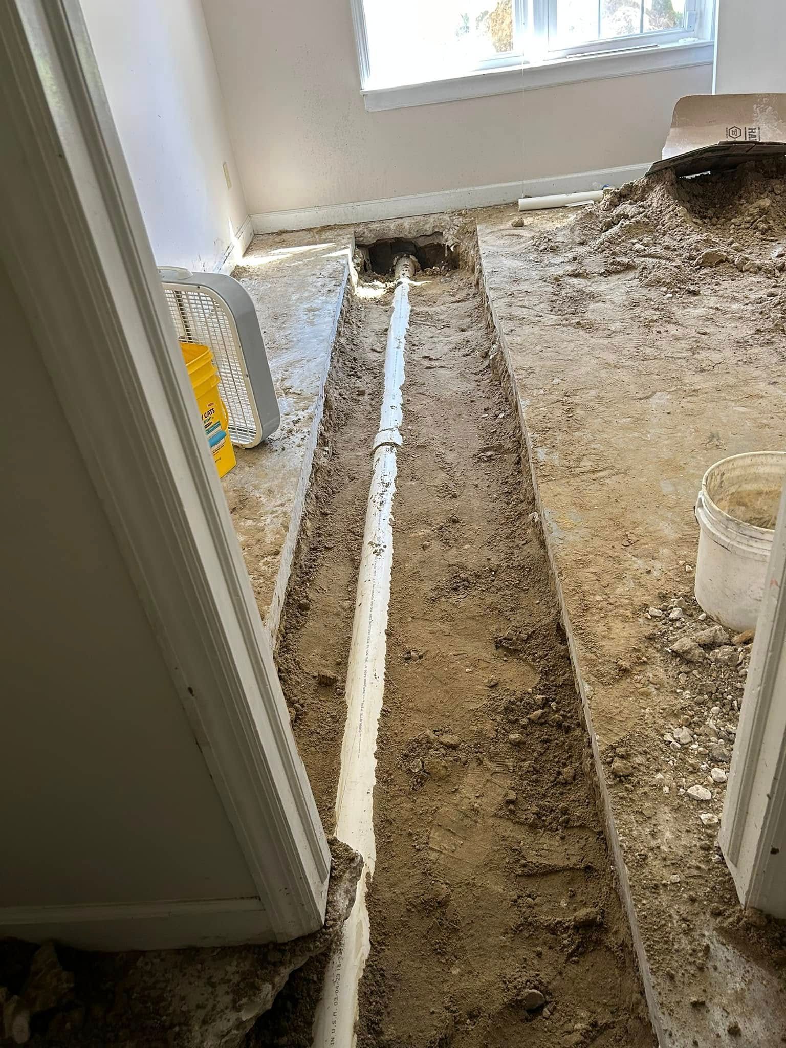 A trench dug through a concrete floor exposing a new white PVC pipe for plumbing repairs in a room.