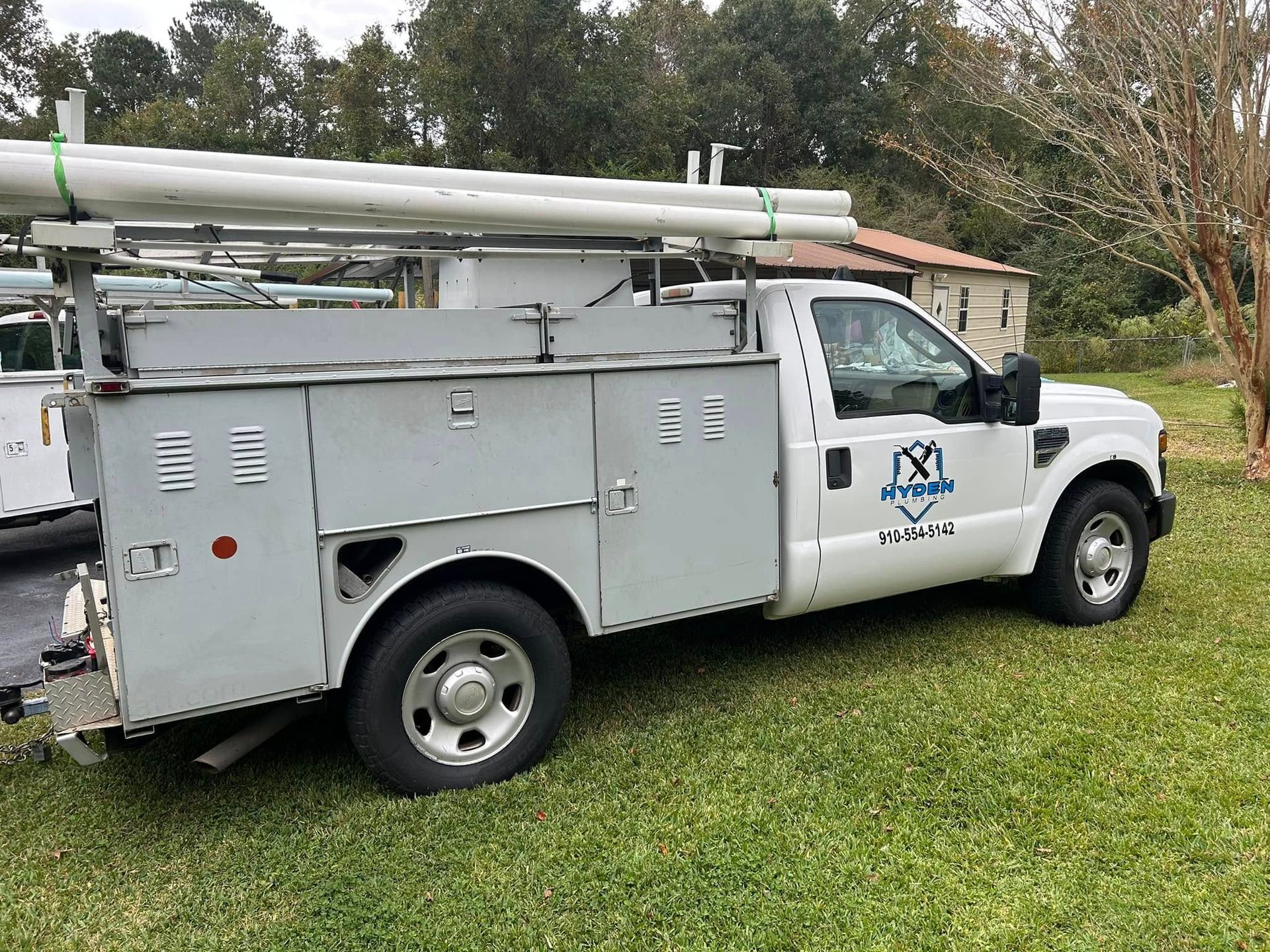 A white utility service truck with storage compartments and a roof rack carrying pipes, parked on a grassy lot.