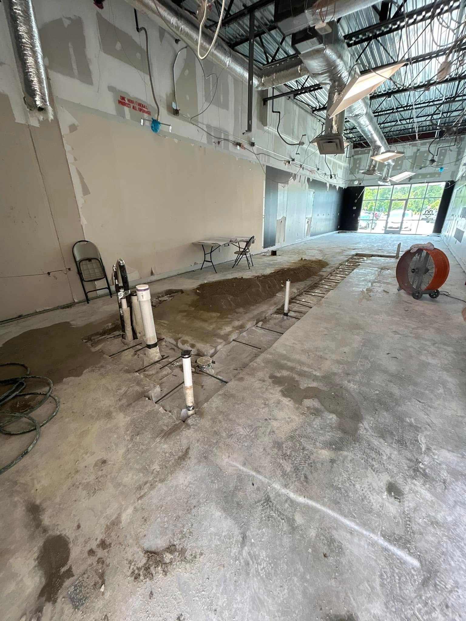 An interior construction site showing an open floor plan with exposed ceiling, concrete floor, and plumbing pipes.