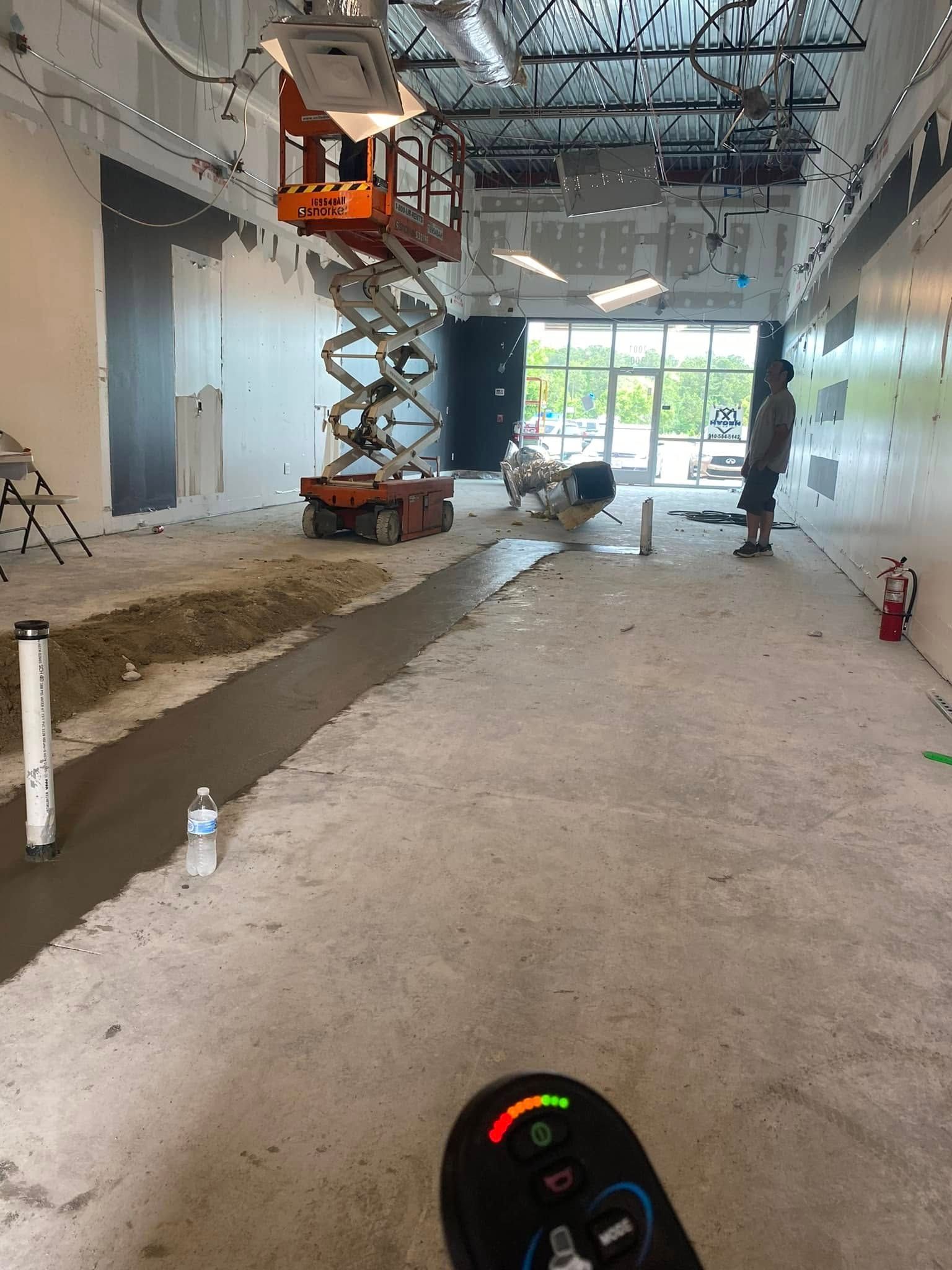 An orange scissor lift stands in a brightly lit, unfinished commercial space with a concrete floor and a strip of wet cement.