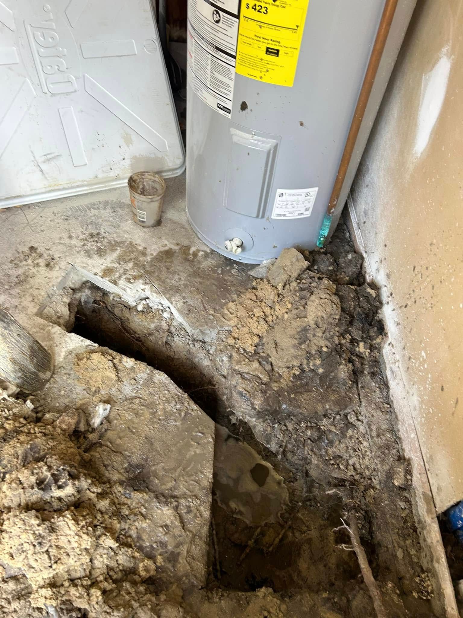 A trench dug into a concrete floor next to a water heater and a wall, revealing dirt and soil underneath.