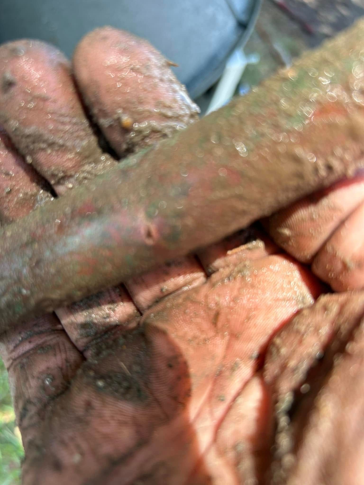 A hand covered in mud holds a muddy, dark-colored cylindrical object or pipe.