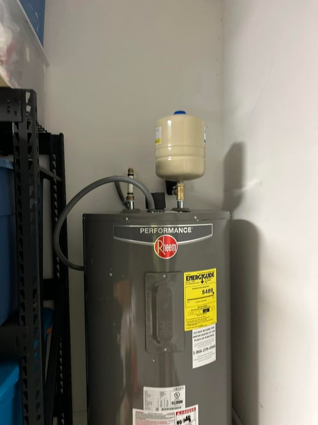 A gray electric water heater with an attached thermal expansion tank stands in a corner next to a storage shelf.