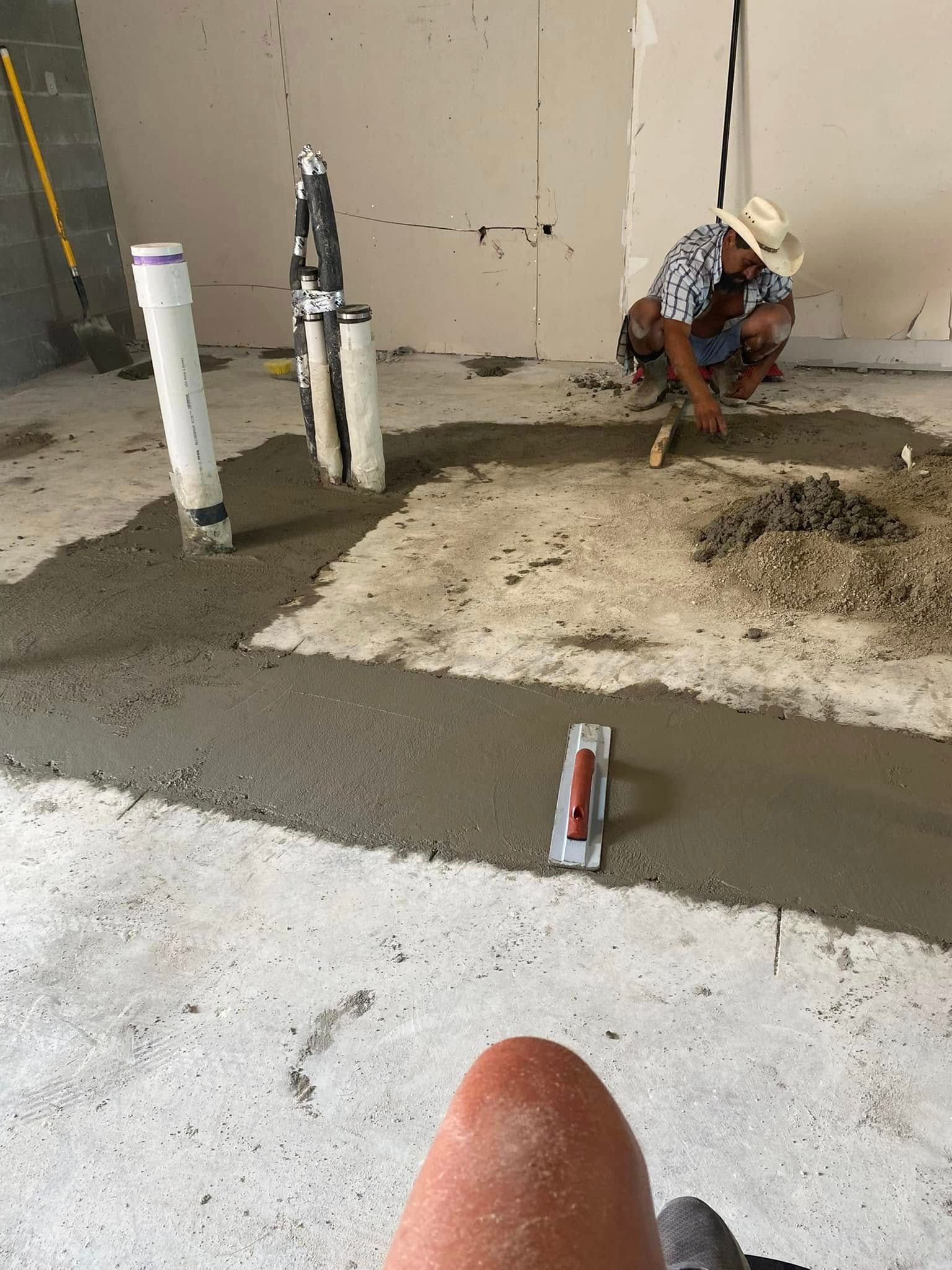 A person in a hat crouches to spread concrete on a construction site floor near plumbing pipes, with a trowel nearby.