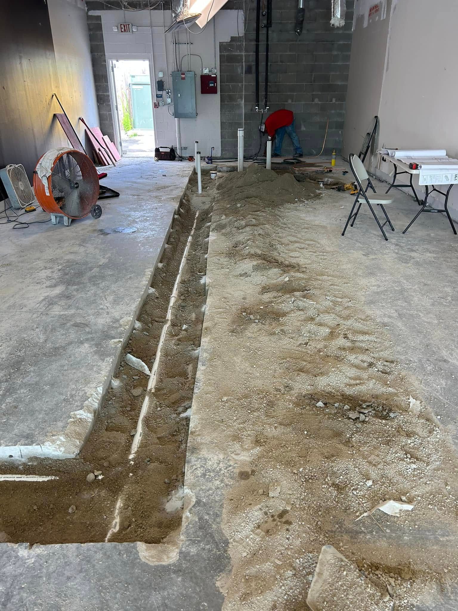 A construction site interior featuring a deep trench cut into a concrete floor, with tools and a worker in the background.