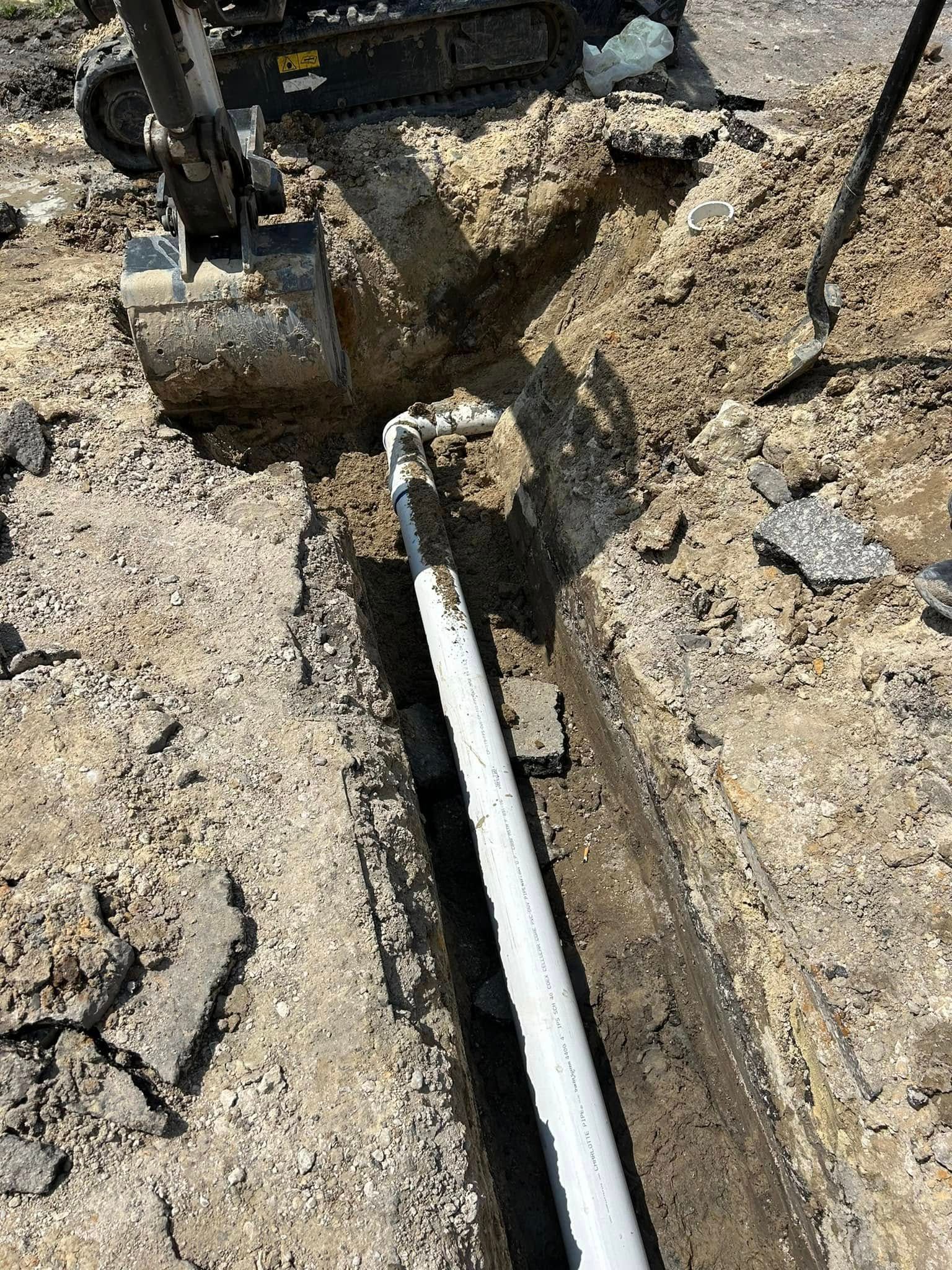 A trench dug into the ground containing a white pipe connected to an elbow joint, with construction equipment nearby.