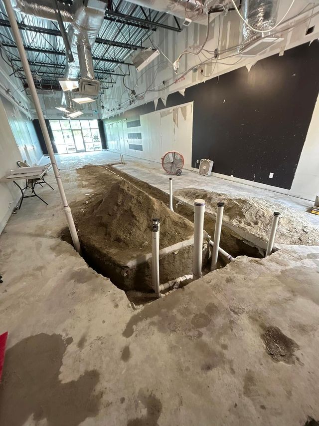 An interior construction site with a long trench cut into a concrete floor, revealing pipes amidst mounds of dirt.