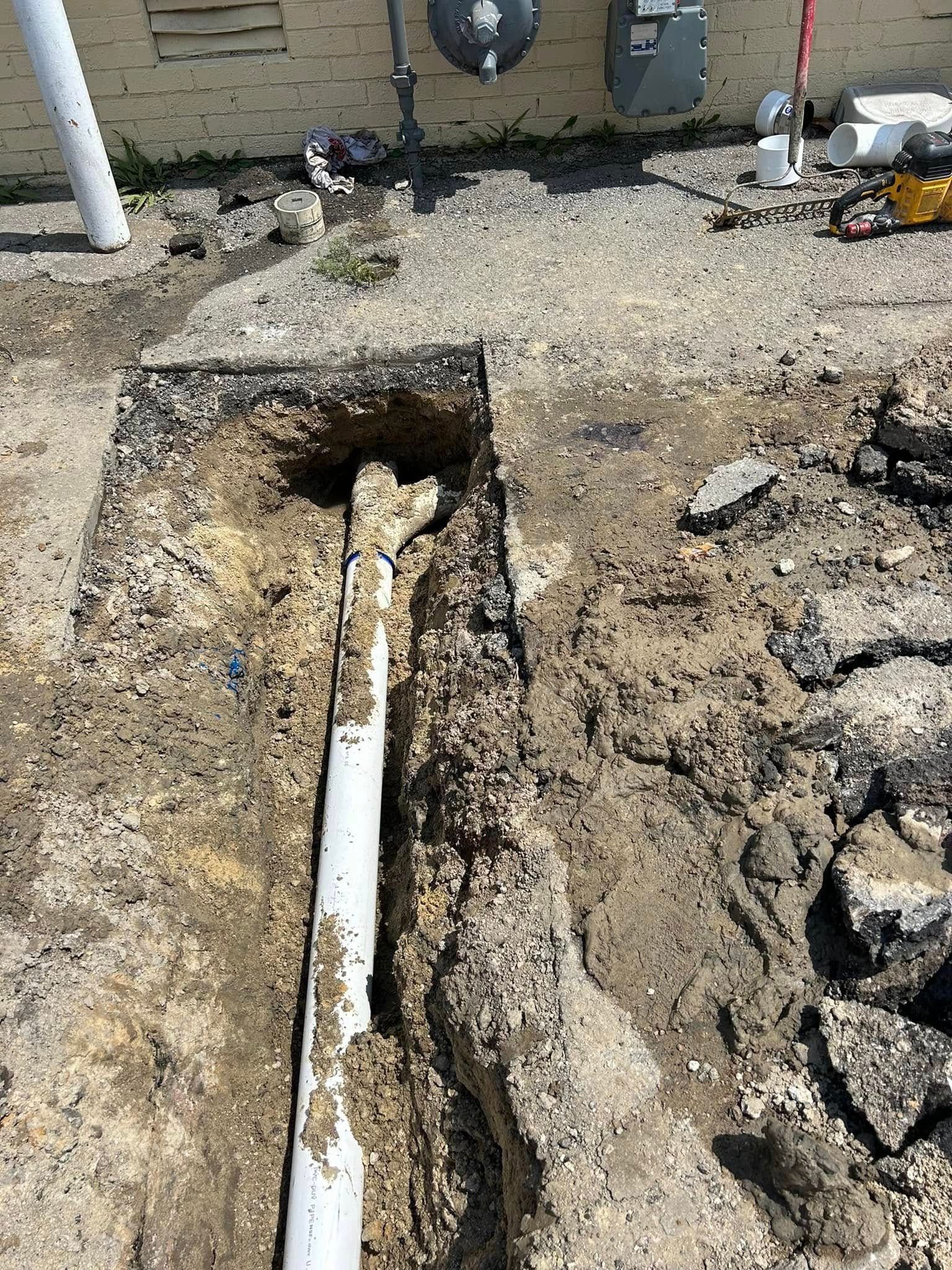A white sewer pipe exposed in a trench dug through dirt and broken asphalt near a building foundation.
