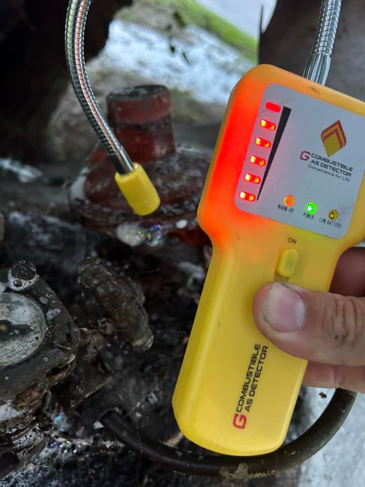 A hand holds a yellow combustible gas detector, showing a high-level alert with red lights near a corroded gas valve.