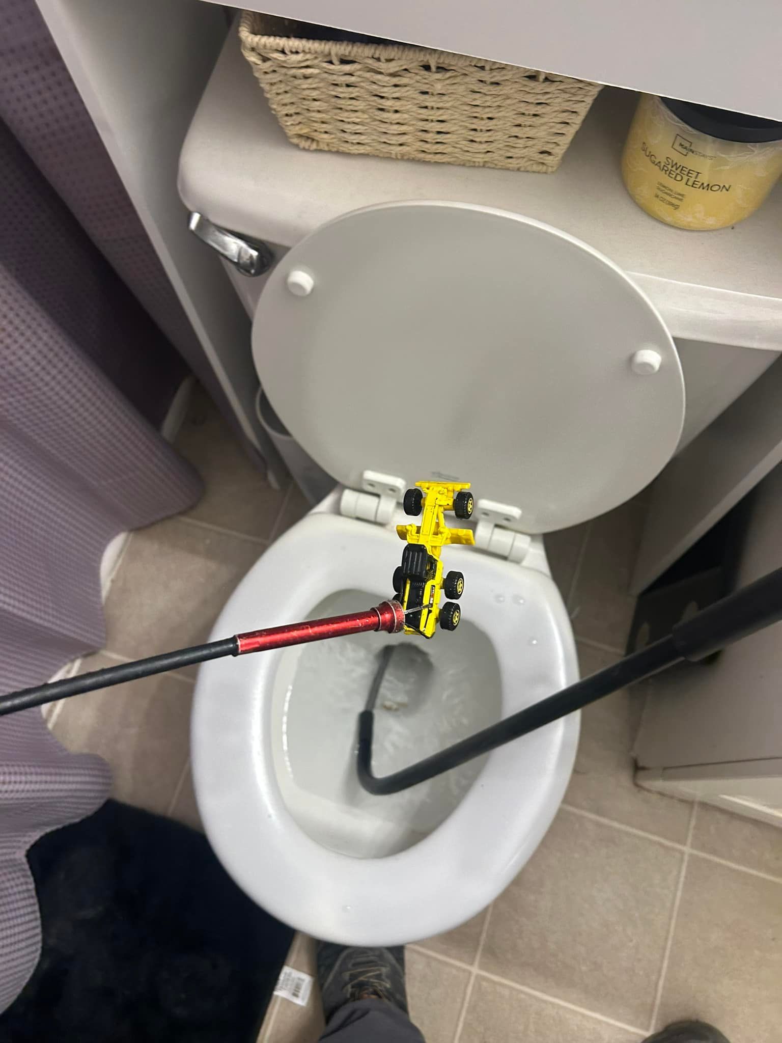 A bright yellow toy truck being retrieved from a toilet bowl with a long, mechanical grabber tool.