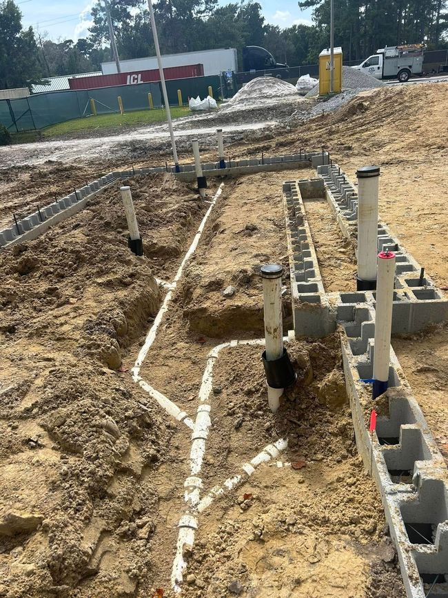 Underground plumbing pipes and vertical vents arranged in a dirt lot at a construction site with concrete block foundations.