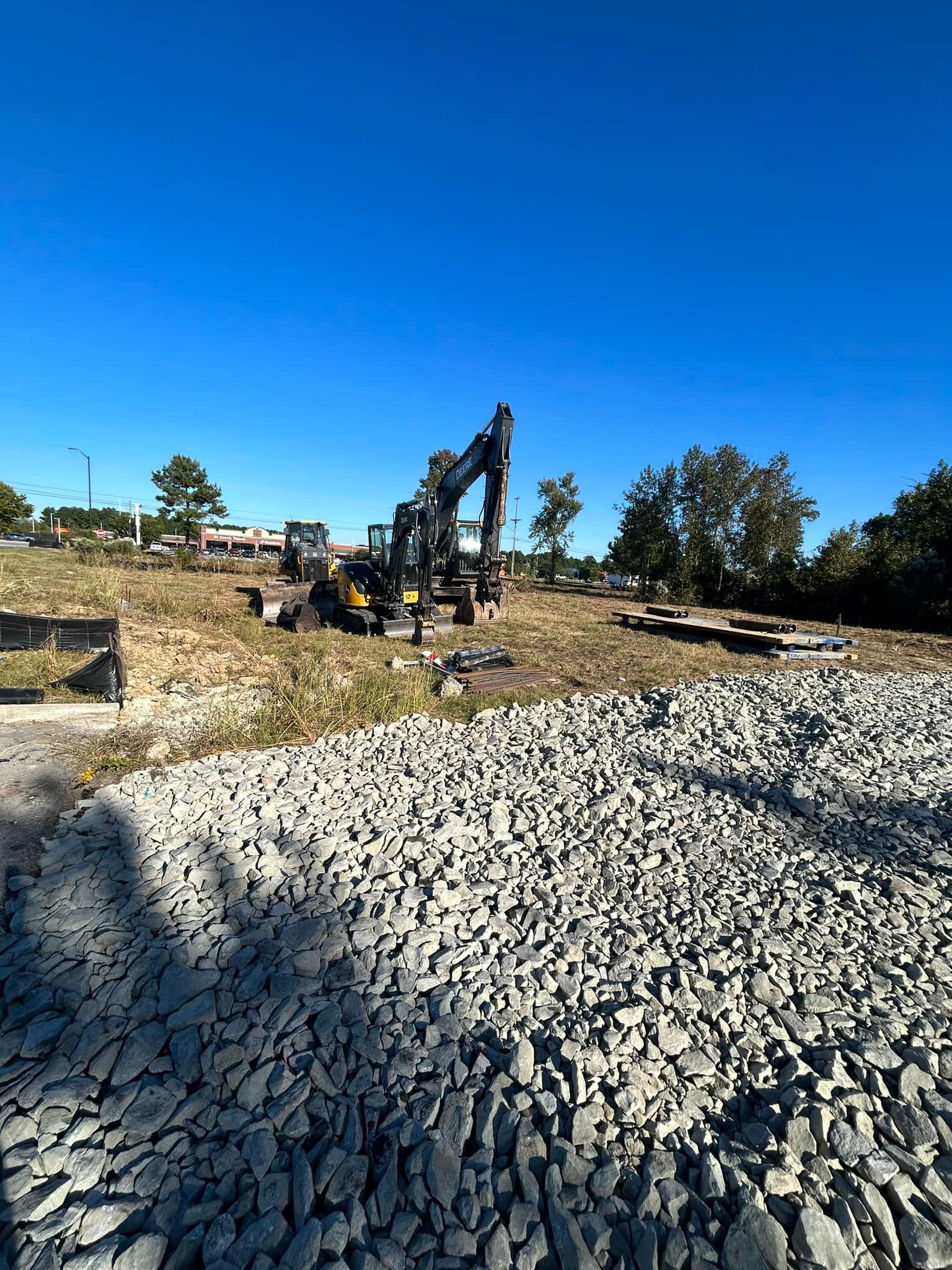 An excavator sits on a dirt construction site covered with a large pile of gray crushed stone under a clear blue sky.