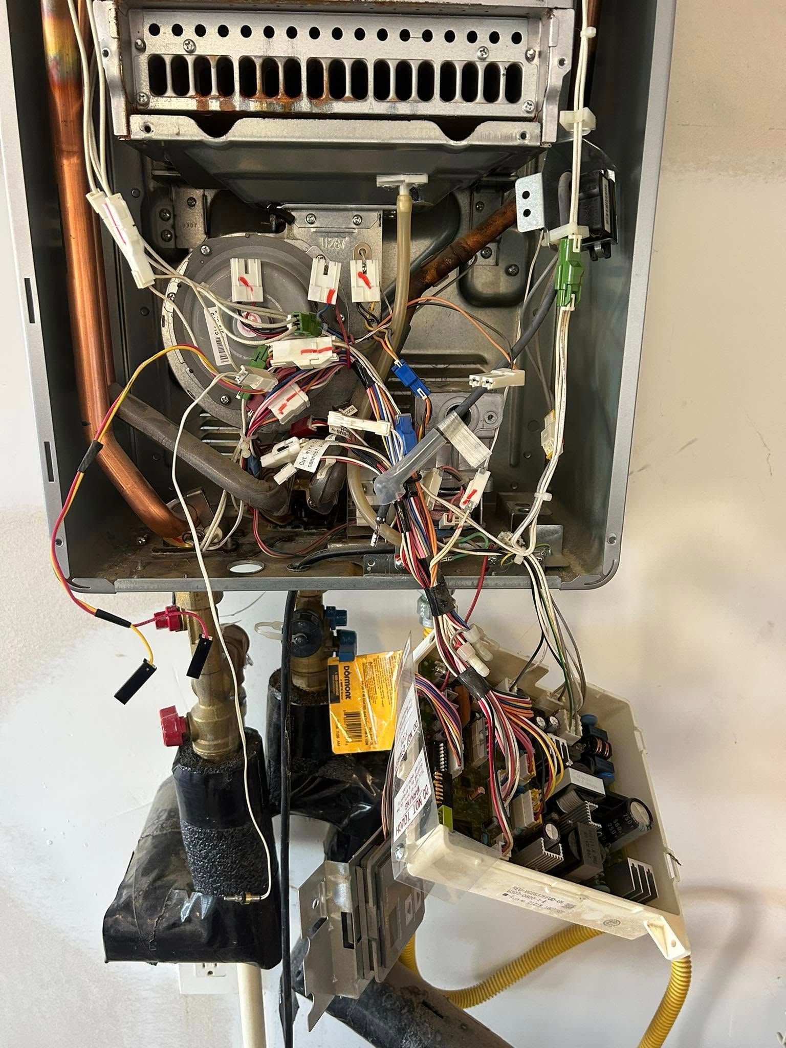 Interior view of an open tankless water heater with exposed wiring, pipes, and internal components mounted on a wall.