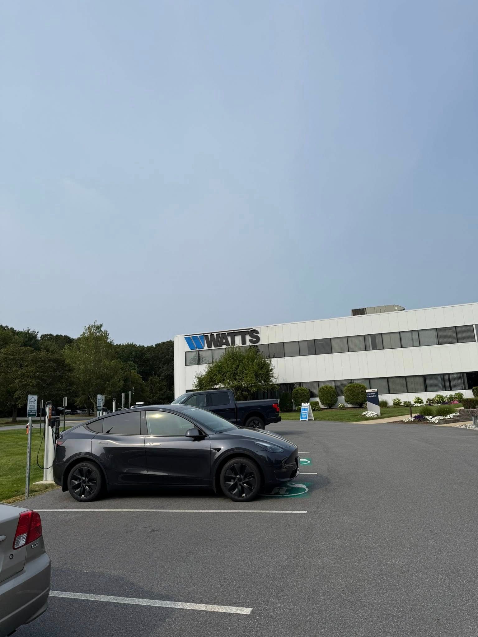 A dark gray Tesla parked at a charging station in front of a white building marked with the name WATTS.