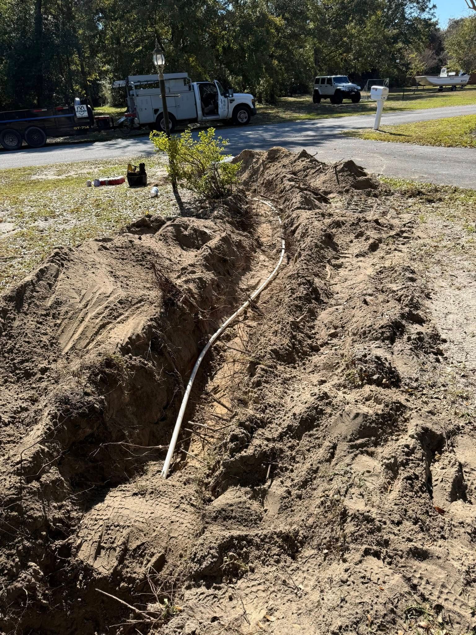 A trench dug into the ground revealing an exposed utility pipe, with a service truck parked on a road in the background.