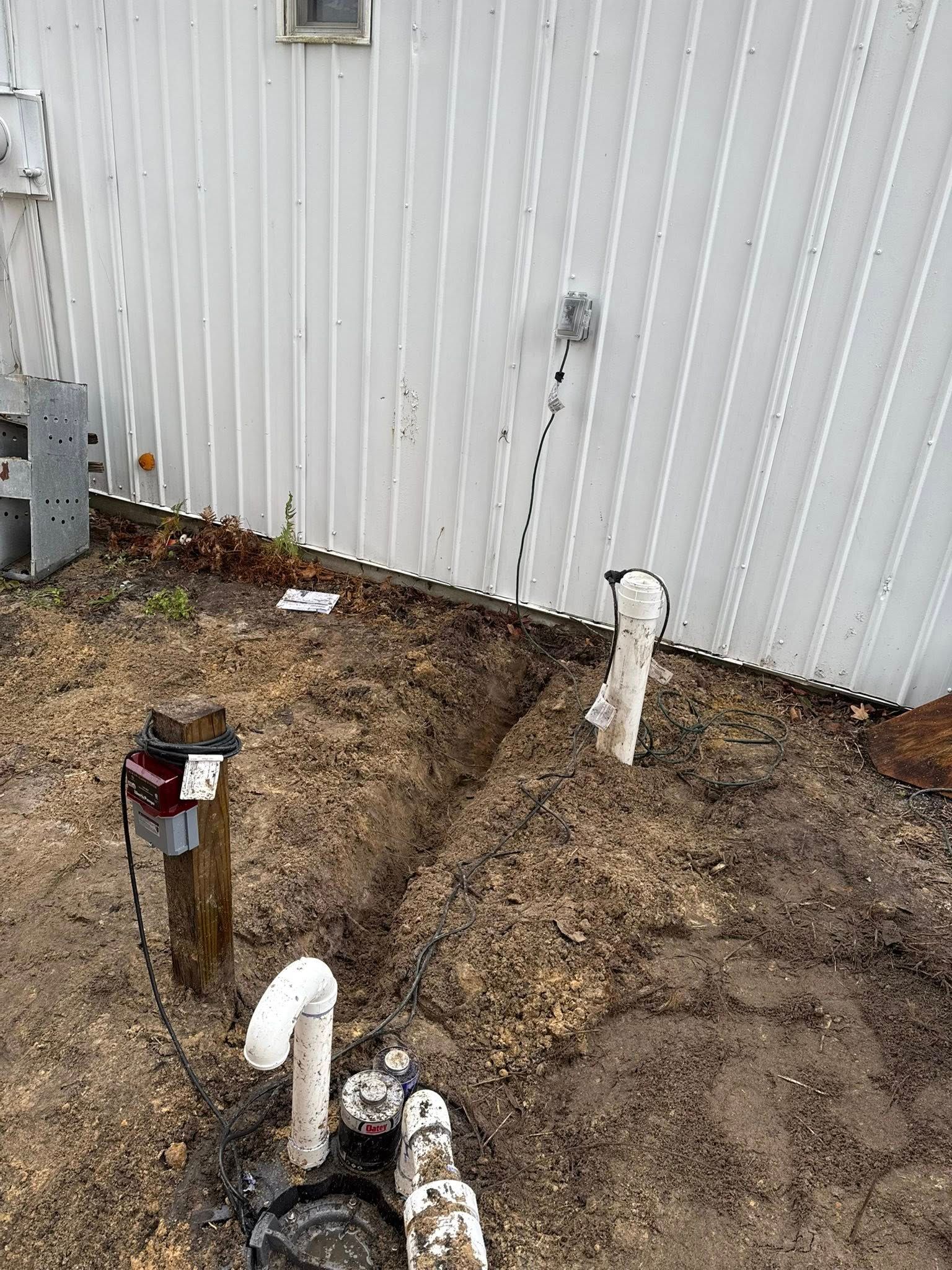 A view of a building exterior with exposed ground, a utility pump, white pipes, and electrical wiring running up the wall.