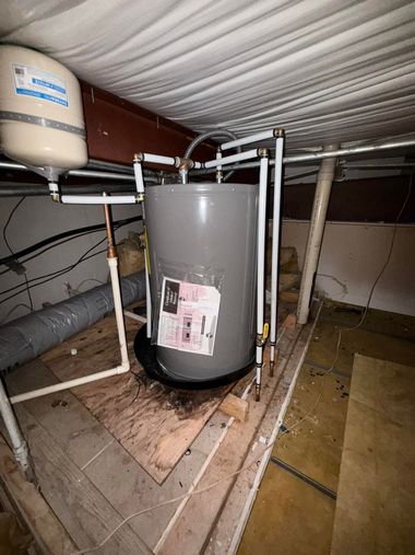 A grey water heater with white piping and an attached expansion tank situated in an attic space.