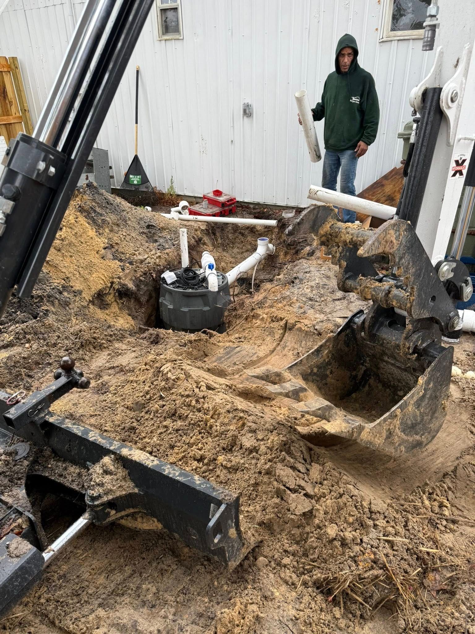 A person in a green hoodie holds a pipe near a construction site featuring a small excavator digging into dirt.