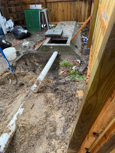 A concrete structure with an open access lid sits in a dirt yard next to a white PVC pipe and a wooden fence.