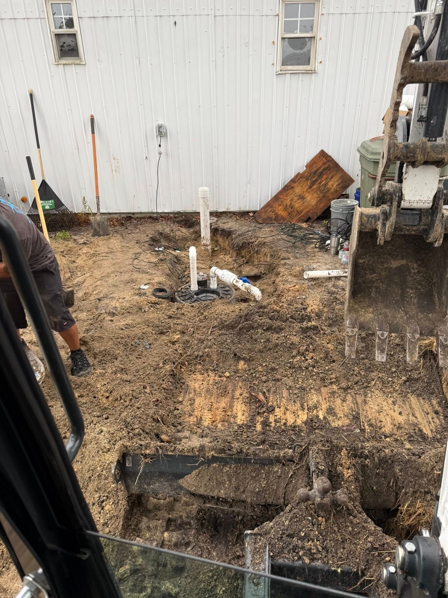 A person operates an excavator to dig near exposed white plumbing pipes in a yard next to a white metal building.