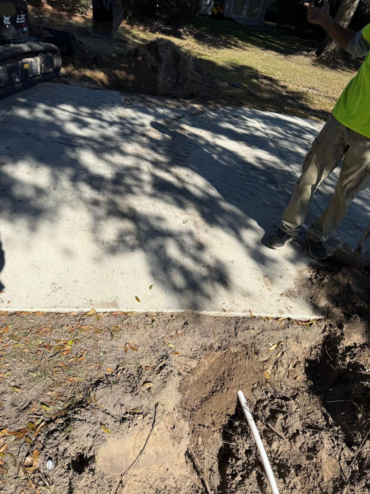 A person in a neon yellow shirt stands next to a freshly poured concrete slab in a dirt area.