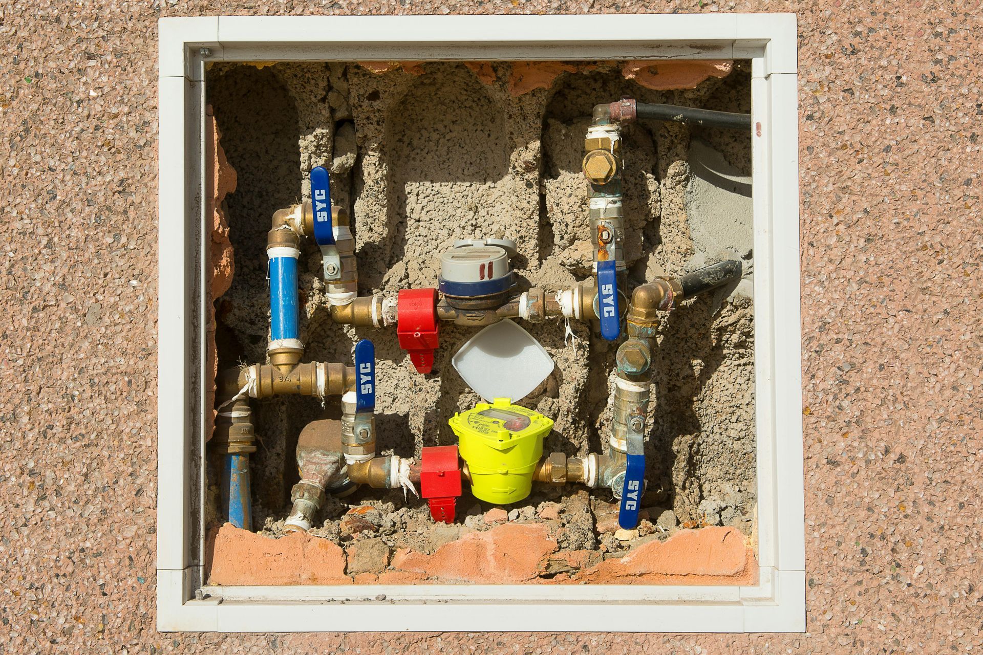 A recessed wall panel showing plumbing pipes with blue-handled valves, red fittings, and two water meters.