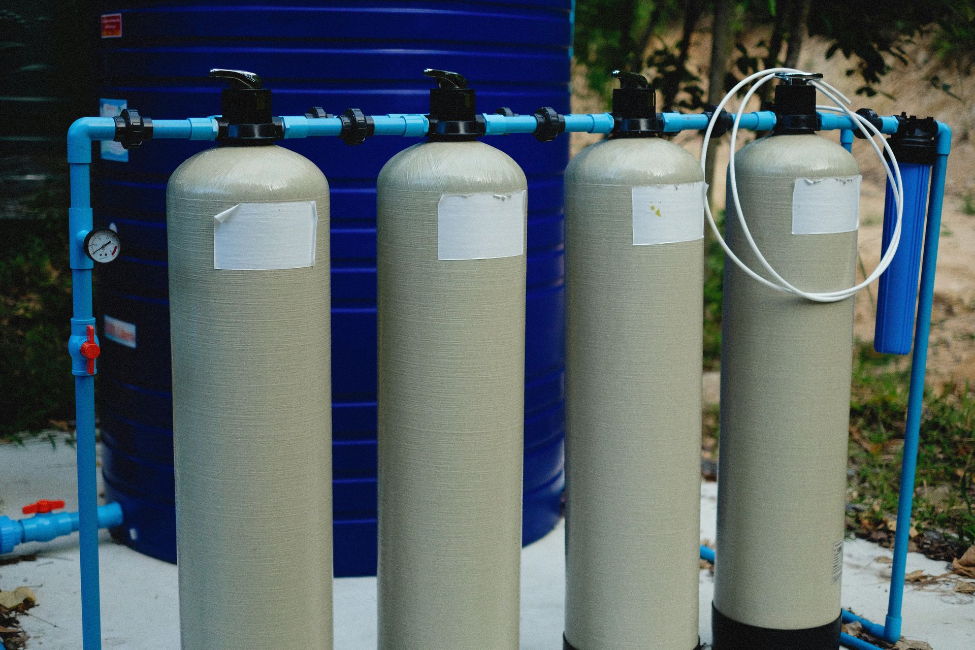 A series of four beige industrial water filtration tanks standing in a row in front of a large blue plastic water tank.