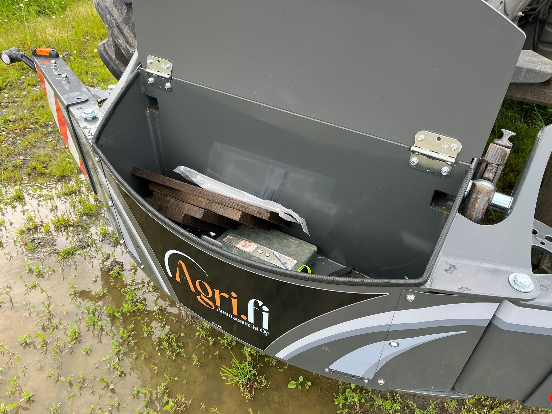 Harmaa traktorin työlaite, osa traktorin etuosaa, varoitussymboleilla ja pienellä oranssilla valolla.