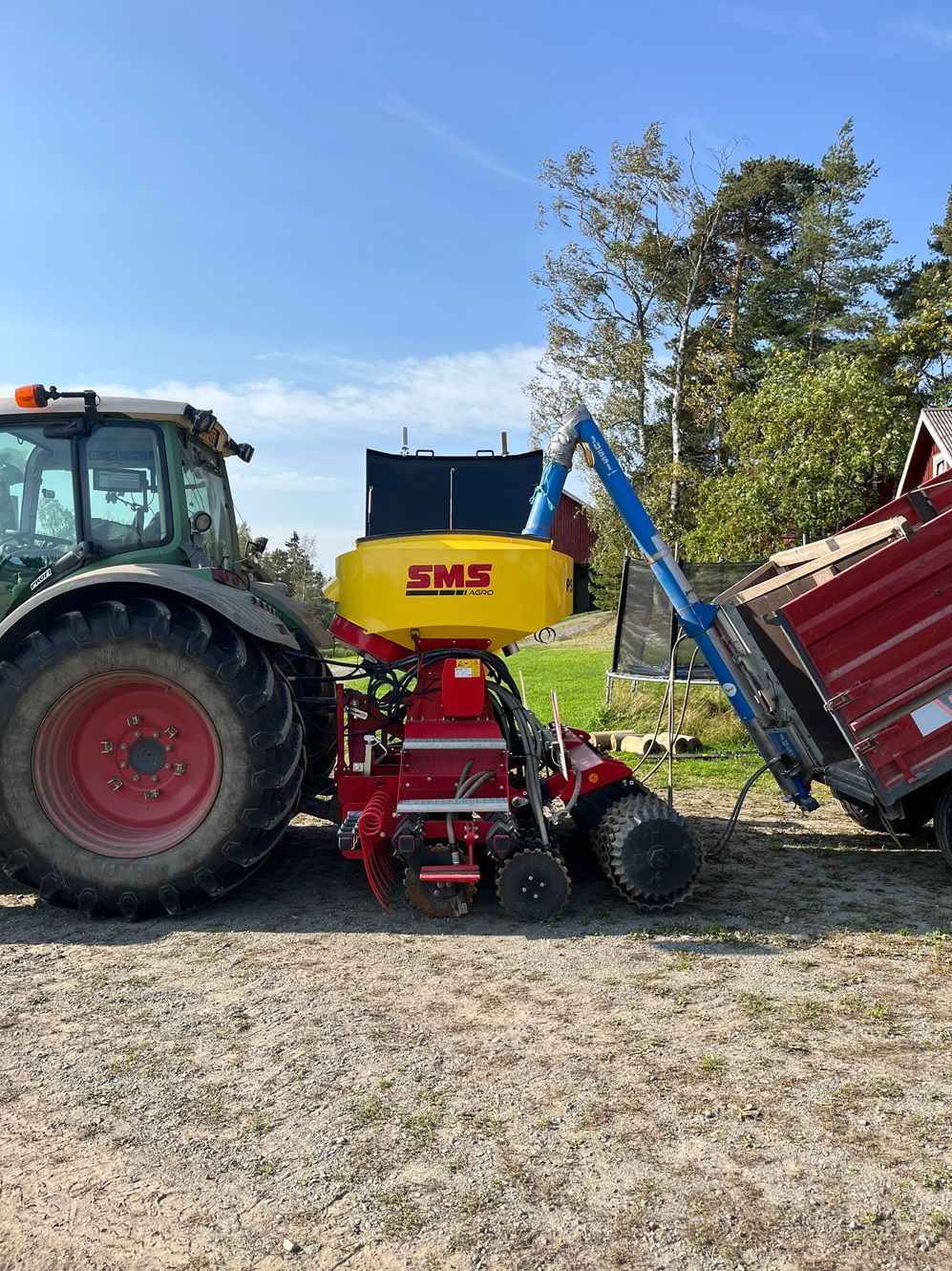Traktori vetää kylvökonetta, johon lastataan viljaa punaisesta kuorma-autosta.