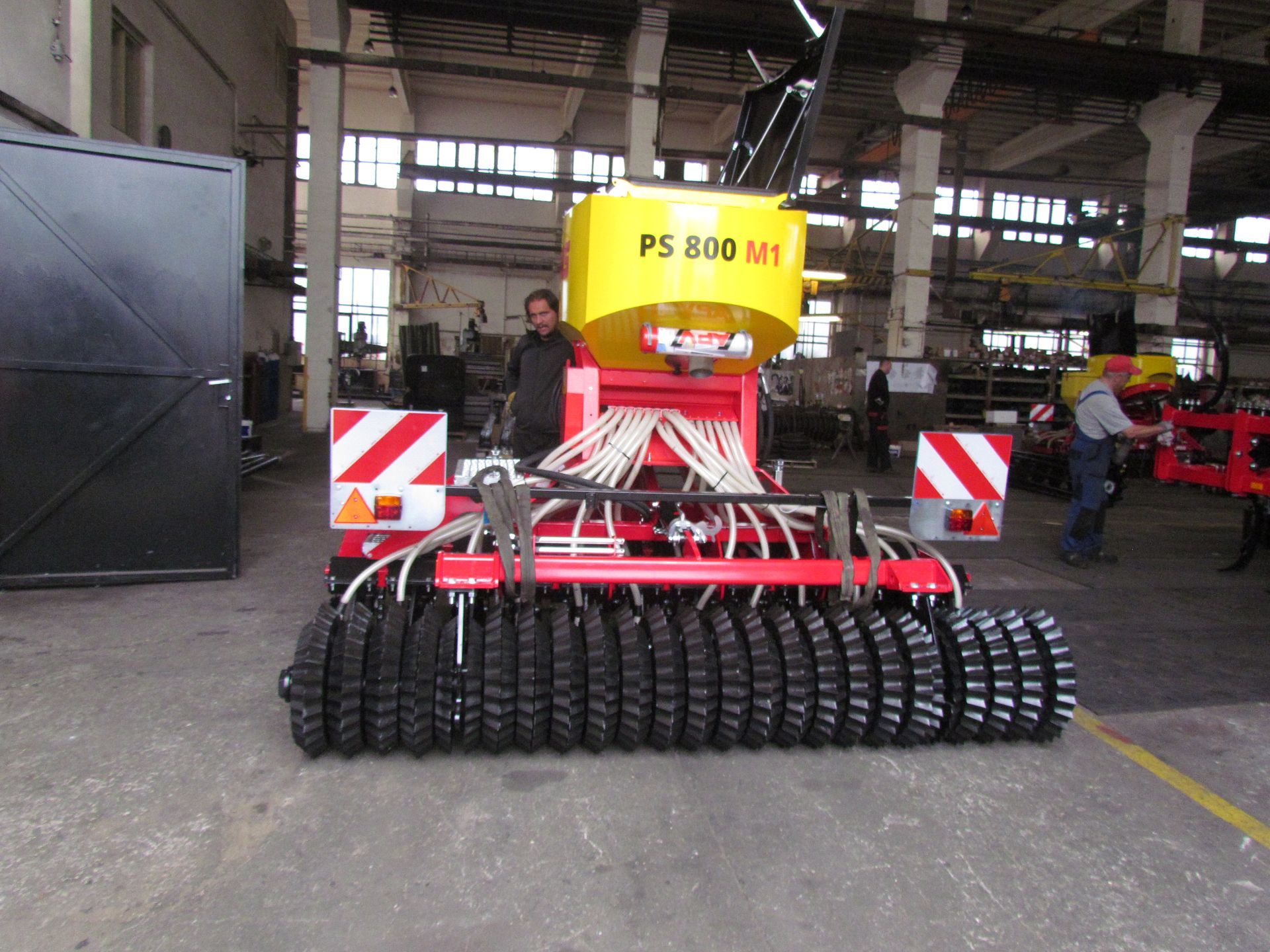 Maatalouden kylvökone, keltainen säiliö, punainen runko, mustat rullat, työntekijöiden kanssa varastossa.